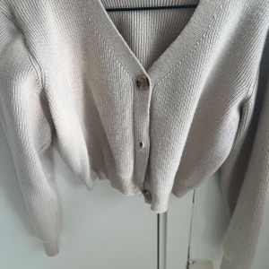 Beige stickad kofta med knappar - Snygg beige stickad kofta med v-ringning och träknappar framtill. Koftan har långa ärmar och ribbad struktur som ger en chill och stilren vibe. Perfekt att slänga på sig när det är lite kyligt ute.