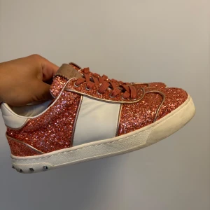 Valentino flycrews glittriga rosa sneakers - Snygga Valentino flycrews sneakers med rosa glitter och vita skinnpaneler. Skorna har klassiska nitar på hälen, matchande rosa snörning och en chunky vit sula. Perfekta för dig som vill sticka ut med en glittrig och exklusiv look.(Alla nitar kvar +låda ingår)