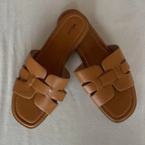 Bruna sandaler från Nelly i skinnimitation - Snygga bruna sandaler från Nelly med breda remmar i skinnimitation och coolt utskuret mönster. Platt sula och fyrkantig tå ger en trendig vibe. Perfekta att glida runt i under varma dagar.