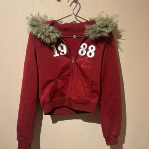Röd hoodiejacka med fuskpäls - Röd hoodiejacka med dragkedja och fluffig fuskpäls runt huvan. Tryck med siffrorna 1988 och texten 'follow heart' på bröstet. Jackan är croppad och har två fickor framtill. Perfekt för dig som gillar en chill och trendig stil. Tyvärr så vet jag inte vart den är ifrån för har haft den ganska länge💓