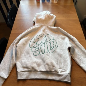Ljusgrå hoodie från LXA STHLM XS - Säljer en ljusgrå hoodie från LXA STHLM i storlek XS. Tröjan har en stor huva, snörning och ett coolt grönt tryck på ryggen med texten LXA STHLM. Perfekt för dig som gillar streetstyle och enkel design.