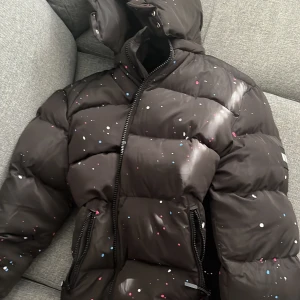 Svart pufferjacka från NVLTY med färgstänk - Svart pufferjacka från NVLTY med coola färgstänk i vitt, rosa och blått. Jackan har huva, dragkedja framtill och två fickor med dragkedja. Perfekt för dig som vill sticka ut i vinter med en unik look. Jackan är i bra skick och köptes förra vintern. Den har storleken XL
