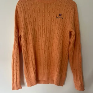 Persikofärgad kabelstickad tröja Bondelid - Kabelstickad tröja från Bondelid i en snygg persikofärg. Tröjan är i 100% bomull, har rund halsringning och långa ärmar. Liten broderad logga på bröstet ger en clean och klassisk vibe. Perfekt för dig som gillar preppy och stilrena plagg.