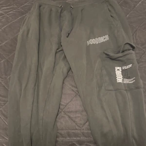 Svarta mjukisbyxor från Hoodrich - Mörka svarta mjukisbyxor från Hoodrich med tryckt logga på benet och en extra ficka med lock. Byxorna har snörning i midjan och är gjorda i 100% bomull för en skön och avslappnad känsla. Perfekta för chill eller streetwear.