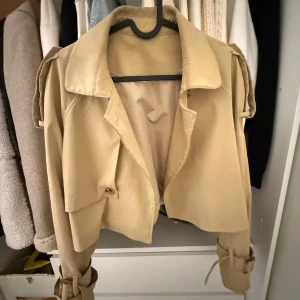 Beige croppad trenchcoat med bälten - Snygg croppad trenchcoat i beige med klassisk krage och bältdetaljer vid ärmsluten. Jackan har axelklaffar och en loose fit som ger en trendig siluett. 
