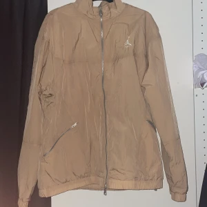 Beige vindjacka från Jordan - Snygg beige vindjacka från Jordan med klassisk logga på bröstet. Jackan har hel dragkedja, två snedställda fickor med dragkedja och meshfoder på insidan. Perfekt för dig som gillar sportig och clean stil. 