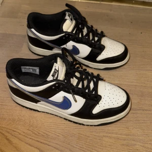 Nike Dunk Low svart - Nike Dunk Low sneakers i svart och vitt med blå swoosh. Klassisk siluett med perforerad tå, svarta snören och vit mellansula. Ovandel i skinn och syntet, låg modell som passar perfekt till streetwear. Snygg kontrast mellan färgerna och ikonisk Nike-logga på plösen.