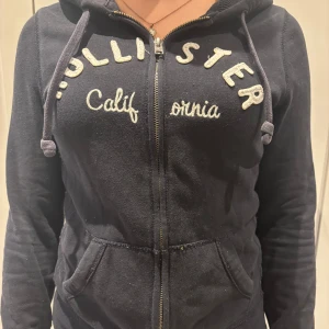 Mörkblå hoodiejacka från Hollister - Mörkblå 2000s hoodiejacka från Hollister med broderad vit logga och text på bröstet. Jackan har dragkedja framtill, huva med snören och två fickor. Perfekt för chill dagar och enkel att matcha med jeans eller leggings.