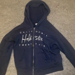 Mörkblå Hollister hoodie med dragkedja XS - Mörkblå hoodie från Hollister i storlek XS med vit broderad text på bröstet. Tröjan har huva med snörning, dragkedja framtill och två fickor. 