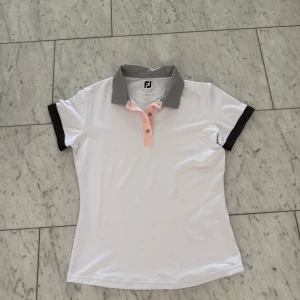 Vit pikétröja från FootJoy, M - Snygg vit pikétröja från FootJoy med grå krage och svarta detaljer på ärmarna. Knappslån är ljusrosa med knappar framtill. Perfekt för golf eller andra sportiga aktiviteter. Tröjan är i mjukt och lätt material som andas.