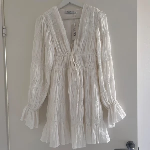 Vit kortklänning från NA-KD med snörning - Superfin vit kortklänning från NA-KD x Moa Mattsson med snygg v-ringning och snörning framtill. Klänningen har långärmade, lätt puffade ärmar och volangdetaljer vid ärmslut och nederkant. 