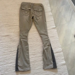 Rock Revival bootcut byxor beige - Säljer ett par snygga  bootcut-byxor från Rock Revival med broderade detaljer och dekorativa bakfickor. Byxorna har låg midja och coola kontrastpaneler i mörkare grått längs benen. Perfekta för dig som gillar statement-jeans med unik design.