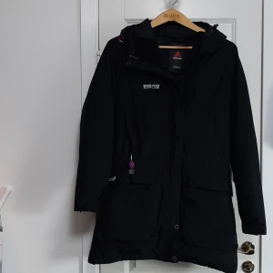 Svart dunparkajacka från 8848 Altitude - Svart dunparkajacka från 8848 Altitude med huva och dragkedja framtill. Jackan har flera fickor, diskreta logodetaljer och en stilren look. Materialet är slitstark polyester som tål väder och vind, perfekt för dig som gillar att vara ute. Snygg och funktionell design.