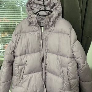 Ljuslila pufferjacka från H&M - Säljer en ljuslila pufferjacka från H&M i storlek M. Jackan har huva, dragkedja framtill och två fickor på sidorna. Perfekt för kalla dagar och riktigt skön att bära. Jackan har en oversized vibe och är superenkel att styla till olika outfits.