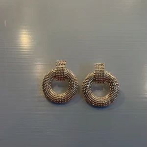 Guldiga cirkelörhängen med struktur - Säljer ett par chunky örhängen i guld med coolt rundad form och detaljerad, prickig struktur på framsidan. De har en modern, statement look och fästs med klassisk stift. Perfekta för att lyfta din outfit med lite extra bling.