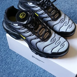 Nike Air Max Plus Black Opti Yellow EU 42 - Nike Air Max Plus sneakers i svart och silver med gula detaljer. Ovandel i mesh med vågformade svarta linjer, svart sula och snörning. Ikonisk Air Max-dämpning och liten gul Swoosh på sidan. Perfekta för dig som gillar en sportig och futuristisk look.