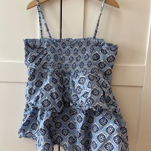 Blå mönstrad topp Gina Tricot - Supersöt blå och svart topp från Gina Tricot med smala axelband och volangdetaljer. Klänningen har ett blommigt mönster och smock upptill som ger en skön passform. Perfekt för varma dagar och sommarhäng med vänner.