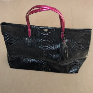 Svart shoppingväska Victoria's Secret - Snygg svart shoppingväska från Victoria's Secret med ormskinnsmönster och långa, glansiga rosa handtag. Perfekt storlek för att få plats med allt du behöver och ger en cool statement-look till din outfit.