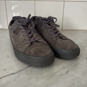 Grå mockasneakers från Yoke - Snygga grå sneakers från Yoke i mjuk mocka med matchande grå snörning och robust svart sula. Skorna har en klassisk rund tå och stilren design som passar till det mesta. Perfekta för dig som gillar en clean och enkel look med lite extra känsla tack vare mockamaterialet. Använda ett par dagar bara då de är för små för mig. Se skick på bilder. Lite trasiga bak vid sömmen vid gummit och ett litet nagg i gummit fram. Köpta i maj 2025. Kvitto finns från Junkyard. Nypris 1169
