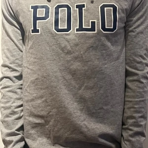 Grå hoodie från Polo Ralph Lauren - Snygg grå hoodie från Polo Ralph Lauren med stor POLO-text i mörkblått på bröstet. Tröjan har huva med snörning och långa ärmar. 