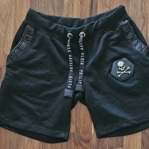 Svarta shorts från Philipp Plein XL - Svarta shorts från Philipp Plein med coolt döskallemärke på ena benet och logotypdetaljer på snörningen och fickorna. Mjukt bomullsmaterial och loose fit, perfekt för chill dagar. Elastisk midja med snörning för skön passform.