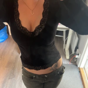 Svart långärmad topp med spets - Svart långärmad topp med djup v-ringning och breda spetsdetaljer längs halsringningen och nederkanten. Sitter tight och har en feminin, elegant look. Perfekt för dig som gillar stilrena och snygga plagg med det lilla extra.