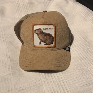 Beige keps Goorin Bros Capybara - Snygg beige keps från Goorin Bros 'The Farm' med patch framtill med en capybara och texten 'LOW KEY'. Kepsen har mesh på sidorna och baksidan för extra ventilation samt justerbar snapback. Skärmen är i brunröd frottéliknande material.