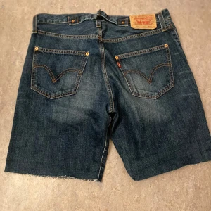 Levi's jeansshorts mörkblå raw cut - Mörkblå jeansshorts från Levi's med råa, avklippta benslut och klassiska femficksdetaljer. Snygga kontrastsömmar och ikonisk läderpatch bak. Perfekta för dig som gillar avslappnade shorts. Storlek W30. 