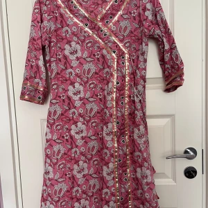 Rosa blommig omlottklänning Glam Roots - Säljer en rosa omlottklänning med blommigt mönster i vitt och svart. Klänningen har guldiga detaljer längs kanterna och trekvartsärm. Materialet är mjuk bomull och modellen är lite loose. Perfekt för dig som gillar färg och mönster!