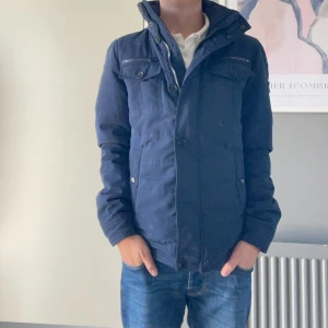 Mörkblå jacka från J.Lindeberg - Snygg mörkblå jacka från J.Lindeberg med hög krage, dragkedja och knappar framtill. Jackan har flera fickor med både dragkedja och knappar, samt diskret logga på insidan. Perfekt för höst och vinter, stilren och enkel att matcha. Passar mig som är 168 fast lite sor i armarna. Så om du är 170 eller någon centimeter till så passar den dig perfekt samt om du är som mig. Bra till hösten kanske och lite på början av våren eller tidig vinter. Inte fast pris!!!!