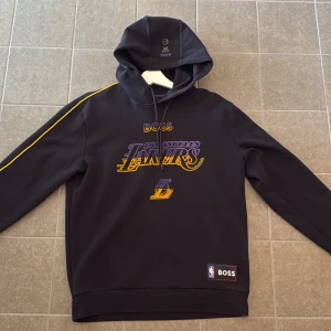 Svart Lakers hoodie från BOSS - Svart hoodie från BOSS x NBA med Los Angeles Lakers-tryck i lila och gult på bröstet. Tröjan har huva med snörning, gula detaljer längs ärmarna och BOSS-märke vid nederkanten. Perfekt för dig som älskar basket och streetwear. Storlek M men passar som S. Kvitto finns. 