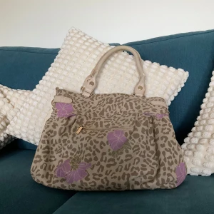 Beige handväska med leopard och lila blommor - Säljer en beige handväska från Essec med leopardmönster och lila blommor. Väskan har ljusa handtag, dragkedja framtill, dekorativ rosett och medföljande liten matchande necessär. Perfekt för dig som gillar unika detaljer och djurmönster.