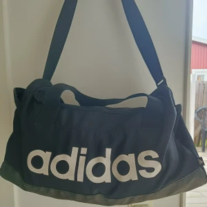 Svart träningsväska från Adidas - Snygg svart träningsväska från Adidas med stort vitt logotryck på sidan. Väskan har justerbar axelrem, bärhandtag, meshficka på sidan och dragkedja upptill. Perfekt för gymmet eller sportaktiviteter. Tillverkad i slitstark polyester.