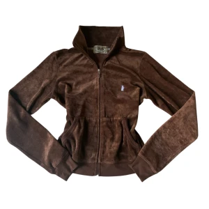 Brun velourjacka från Juicy Couture - Säljer en brun velourjacka från Juicy Couture med dragkedja framtill och hög krage. Jackan har två fickor och en liten broderad logga på bröstet. Perfekt för dig som gillar den klassiska Y2K-stilen och vill ha något mjukt och bekvämt.