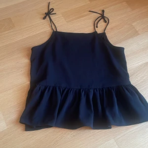 Svart volangtop från H&M med knyt - Svart linnetopp från H&M med tunna axelband. Toppen har en rak halsringning och är helt slät utan mönster. 🖤🖤