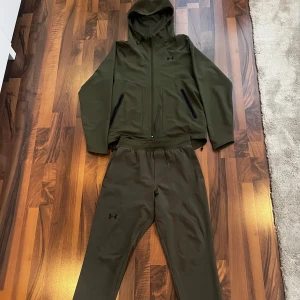 Under Armour grön träningsset med huva - Snyggt träningsset från Under Armour i mörkgrön färg. Setet består av en hoodie med dragkedja, huva och svarta detaljer samt matchande byxor med resår och snörning i midjan. Perfekt för träning eller chill. Materialet är mjukt och stretchigt syntet.
