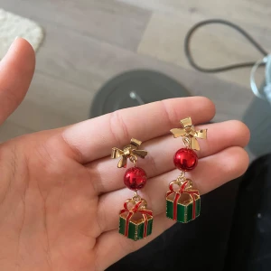 Jul örhängen - Superfina örhängen med guldiga rosetter, röda glittriga kulor och hängande gröna paket med röda band. Perfekta för att maxa julstämningen och sticka ut på alla vinterhäng. Färgerna är guld, röd och grön, och detaljerna är riktigt festliga.