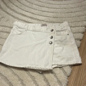 Vit jeanskjol med shorts från ONLY - Supersnygg vit jeanskjol från ONLY med inbyggda shorts under. Kjolen har tre silvriga knappar framtill, bälteshällor och klassiska fickor både fram och bak. Perfekt för dig som vill ha en cool och bekväm look i sommar.