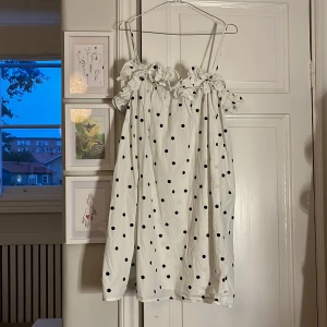 Vit prickig kortklänning från H&M - Supersöt vit kortklänning från H&M med svarta prickar och volangdetaljer vid axlarna. Klänningen har tunna axelband och en luftig passform. Inte använd någon gång men köpt secondhand!