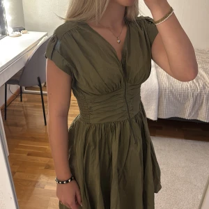 Olivgrön kort klänning med dragkedja - Snygg olivgrön klänning med v-ringning och dragkedja framtill. Modellen är kort med markerad midja och lätt puffade axlar med knappdetalj. Materialet är tunt och har ett lite glansigt utseende, perfekt för dig som gillar en avslappnad men chic stil.