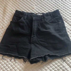 Svarta jeansshorts från Lindex - Svarta jeansshorts från Lindex med hög midja och råa, fransiga benslut. Klassisk femficksmodell med bälteshällor och dragkedja. Perfekta för en chill sommarlook och lätta att matcha med allt.