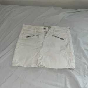 Vit jeanskjol med dragkedjor - Snygg vit jeanskjol från Blend med två dragkedjor framtill och klassiska fickor både fram och bak. Kjolen har hällor för bälte och en rak, kort passform som ger en fräsch och stilren look. Perfekt för dig som gillar enkel och cool stil.