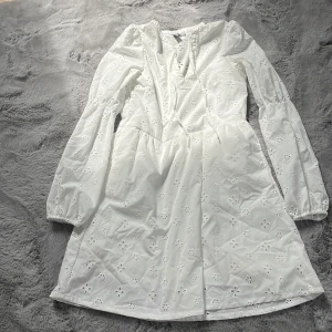 Vit klänning med broderade blommor från H&M Divided - Söt vit klänning från H&M Divided med broderade blommor och långa puffärmar. Klänningen har omlott-skärning upptill och resår vid ärmsluten. Perfekt för vår och sommar!