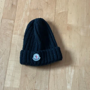 moncler mössa - Svart ribbstickad mössa från Moncler med uppvikt kant och klassisk Moncler-logga framtill. Perfekt accessoar för kyliga dagar och enkel att matcha med olika outfits.