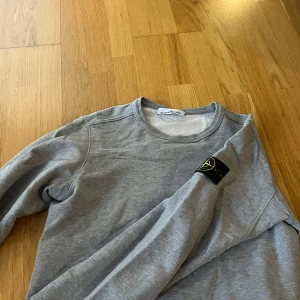Grå sweatshirt från Stone Island - Säljer en stilren grå sweatshirt från Stone Island med klassisk rund hals och logotyp-patch på ärmen. Tröjan har en enkel design och är perfekt för en avslappnad look. Passar dig som gillar streetwear och snygga basplagg. Storlek M