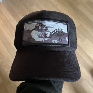 Svart keps med vintage biltryck - Svart keps med ett stort tygmärke framtill som visar ett svartvitt motiv av en person som kör en gammal bil. Kepsen har böjd skärm och justerbar snapback baktill i mesh. Perfekt för dig som gillar retrostil och bilar.