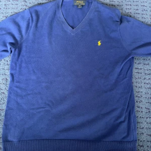 Blå v-ringad tröja från Polo Ralph Lauren - Säljer en klassisk blå v-ringad tröja från Polo Ralph Lauren med gul broderad logga på bröstet. Tröjan har långa ärmar och ribbade muddar vid ärmslut och nederkant. Perfekt för en stilren look.