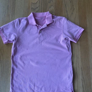 Pikétröja Uniqlo - Säljer en ljuslila pikétröja från Uniqlo med klassisk krage och två knappar framtill. Tröjan är kortärmad och tillverkad i mjuk bomull, perfekt för en avslappnad stil.