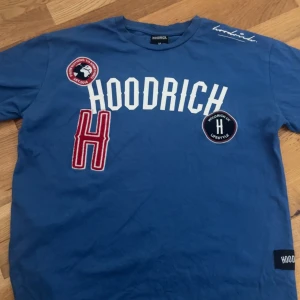 Blå t-shirt från Hoodrich med tryck - Säljer en blå t-shirt från Hoodrich med stora vita tryck och patchar på bröstet. T-shirten har korta ärmar och rund hals. Perfekt för dig som gillar streetwear och vill sticka ut med snygga detaljer.
