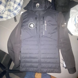 Svart JR Antarctic Hybrid Hood jacka från Sail Racing - Säljer en svart Sail Racing JR Antarctic Hybrid Hood jacka med huva och dragkedja. Jackan har broderade loggor på bröstet och ärmen samt quiltade partier framtill. Perfekt för kyliga dagar. Fick den i present och passar perfekt för dig som är 170-183 lång, säljer den för jag ej passar i den 